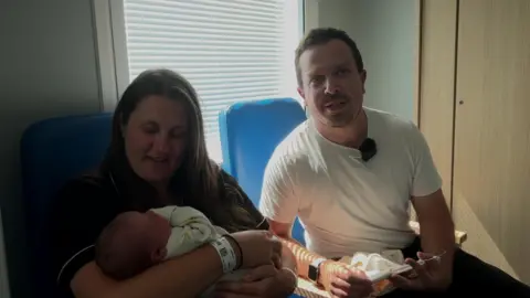 A man and woman sit in a dimly-lit room in front of a window with a Venetian blind. The man is looking directly at the camera while the woman, who is holding a baby in her arms, looks down at the child. 