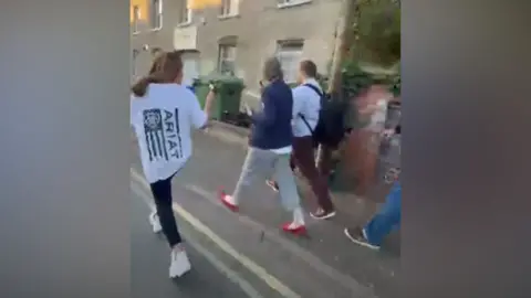 Facebook A woman holds her phone while videoing a man and a woman walking along the street, in a blurry screenshot from mobile phone footage.