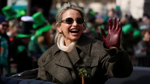 REUTERS/Cathal McNaughton Vogue Williams is in an open top car and waving at a large crowd. She has shamrock pinned to her checked coat. She's wearing a cream turtle neck jumper and maroon gloves. Some people in the background have leprechaun hats on. 