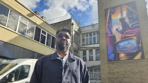 Aisha Iqbal/BBC Artist Mohammed Ali Aerosol is pictured outside a building bearing a mural which shows a car with the wing mirror projecting an image of a building