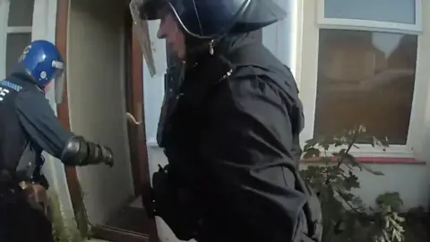A police raid in Middlecroft Lane, 21.8.24. Two officers wearing protective helmets and face guards are moving near a property's open front door.