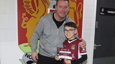 Contributed Kevin puts his arm around Ezra and they both smile at the camera. They are inside with a large sign of the Northampton logo behind them, against white brick walls. Kevin wears a grey hoodie and black trousers.