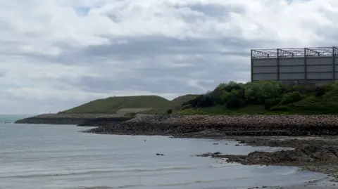 A photo of La Collette's Eastern Headland as viewed from Harve de Pas. In the foreground is the sea, and beyond that rocks piled up to make a mound, with a grassy hill on top to the left of the picture, and a big metal building to the right of the picture.