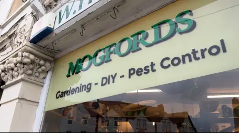 The outside of a shop window. The sign on the window reads: "Mogfords Gardening - DIY - Pest Control"
