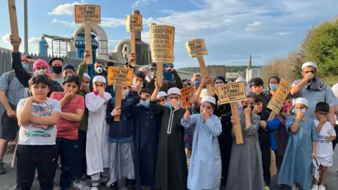 A large group of children, with a few adults, stand posing with placards saying 'end the stink'. Most of them also hold their noses in disgust. Behind them is a factory, with large, shiny metal pipes visible.