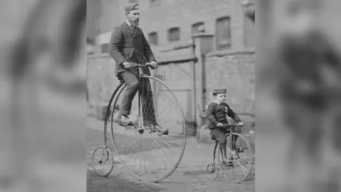 Leeds Museums and Galleries A black and white picture of a man and boy riding matching penny farthing bikes on a cobbled street. They are both dressed in suits and a hat.