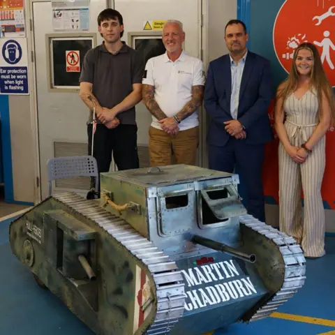 Hull 4 Heroes Three men and a woman are smiling next to a soapbox kart designed as a green tank with the name Martin Chadburn inscribed on the front in white lettering.