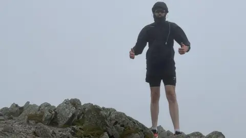 Summit for Grange Toby stands on a mountain top with rocks, wearing shorts and a jacket. You can hardly see his face as he has his hood up and sunglasses on. 
