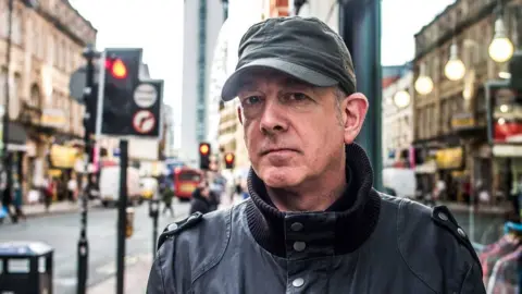Dave Haslam, a man wearing a black coat and a dark-coloured baseball cap, is stood near some traffic lights in a city centre.
