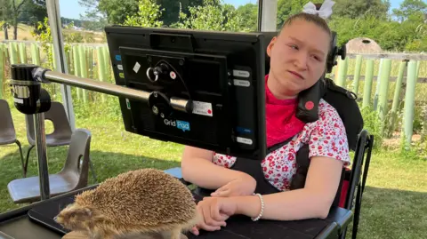 Isabella is sat in a wheelchair next to a table which has a taxidermi hedgehog on it. She has brown hair tied up with a white bow and is wearing a white and red floral top.