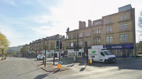 A Google Streetview image of Briggate, showing a white van pulled up at a set of traffic lights. A row of three storey-buildings line the right hand side of the road. There are trees on the left.