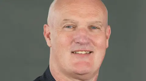 BBC Kenny Mcintyre, who has a bald head, is looking into the camera and smiling.