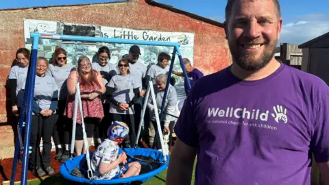 Image of a man with a beard wearing a purple charity tee shirt who is smiling at the camera. In the background can be seen a group of people in grey tops against a wall and a boy in a blue swing with his head turned away from the camera.