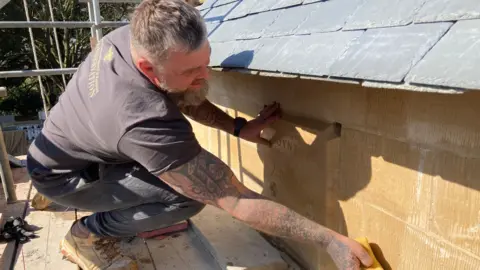 Scott Dalton / BBC A tattooed man in a t- shirt is crouching on scaffolding and sponging down sandstone bricks on the church. You can faintly see one of the bricks has a carving in it saying High Toynton - 2025.