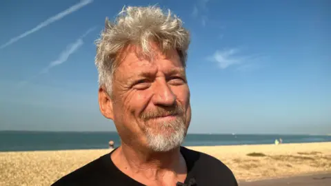 Mark is wearing a black t-shirt and has grey hair and a grey beard. He is standing on the beach facing away from the sea.