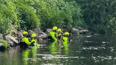 BBC/Phil Bodmer A number of people wearing waterproof high-vis clothing stand in a river up to their chests
