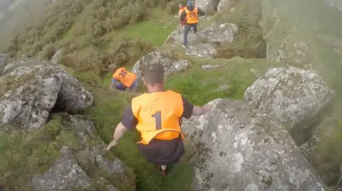 Jim Thurston A group of people clamber over a set of large rocks on Dartmoor. They are wearing orange bibs, with the numbers 1, 38 and 25 written on them. 