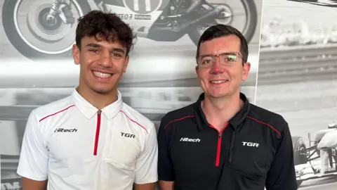 A young man in a white polo shirt and an older man in a black version of the same shirt stand side-by-side with broad smiles.