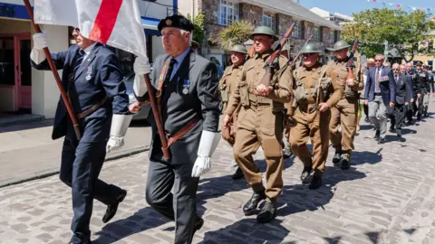 Two men in suits with war military medals over their left breast carry a large red and white flag with soldiers in historic military uniform following behind them. Behind the soldiers is a group of people in smart dress who also have military medals over their left breasts. 