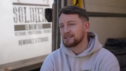 Lewis Kendrick, wearing a light grey hooded jumper, sitting in the back of a removal van.