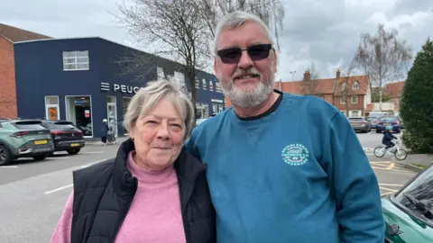 A couple in a car park, the woman has grey/blonde hair and wears a pink jumper, the man wears a blue jumper, sunglasses and has grey hair.