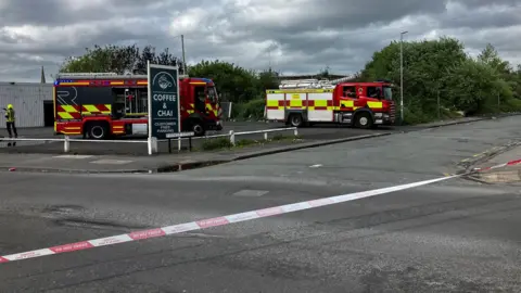 Two fire trucks in a parking lot on a cloudy day. The parking lot is at the junction of two roads. One of the roads is blocked off with red and white tape.