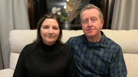 Alan and Debbie Mead sitting on their sofa. Alan is wearing a green and blue checked shirt and has grey hair. Debbie is wearing a black turtle neck jumper.