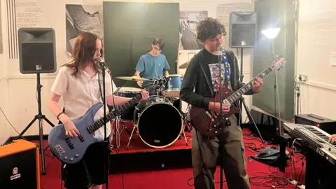 A teenage girl and boy playing guitars and singing into microphones. There is a teenage boy playing drums behind them. 