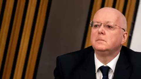 Peter Murrell in a black suit, white shirt and black tie. He is wearing glasses and looks very solemn. 