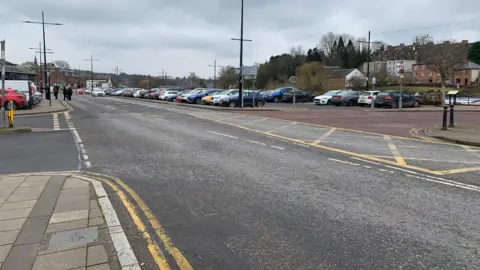 A busy car park on the Whitesands area in Dumfries with numerous cars parked up next to a quiet road with houses in the background across the river