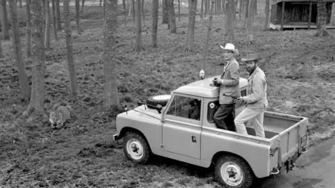 PA Media The 6th Marquess of Bath stands on his Land Rover Defender backed by an armed warden, watching a lion at his game reserve in Longleat. They're both wearing wide brimmed hats and jackets, and are looking at the camera