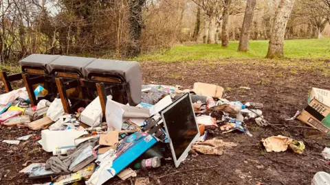 A pile of rubbish in a field including a broken sofa, a computer monitor and other rubbish surrounded by muddy ground.