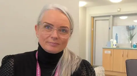 Claire Johnson has long silver hair in a ponytail sitting in an office, wearing a black polo necked jumper and  "workplace champion" lanyard and glasses looking at the camera.