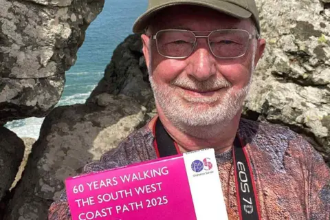 Rick Cornwall A man with a brown cap and silver glasses smiling. He's along the coast path and has a camera round his neck and a pinky T-shirt. He's holding an OS map which says 60 years walking the South West Coast Path 2025