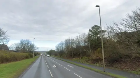 Three lane South Road, with grass, hedges and trees on each side. There are vehicles in the distance, and white road markings. 