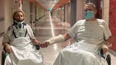 Mante Linkus Saule Linkus, in a neck brace, holds hands with husband Darius Linkus as the pair sit in wheelchairs in a hospital corridor