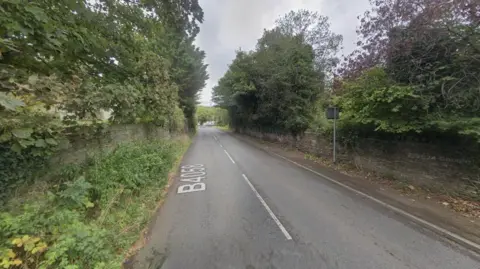 A stretch of road with stone walls, trees and bushes either side of it. There are no vehicles on the road.