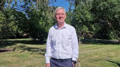 BBC Tim Oliver looking at the camera. He is wearing a grey shirt and blue trousers. Grass and trees in the background.