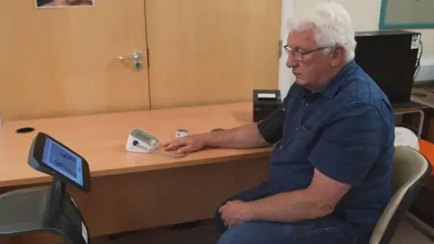 A man seated in a clinic or office consultation room using a digital health monitoring device. A blood pressure cuff is attached to one arm, with the device resting on a desk. A touchscreen unit stands nearby, and computer monitors are visible in the background.
