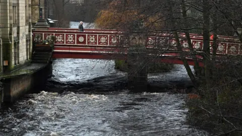 A red footbridge runs over a tempestuous-looking river while a pedestrian walks across. On the riverbank are bear, leafless trees.