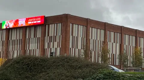 BBC/Federica Bedendo A general view of the Sands Centre in Carlisle taken from the pedestrian underpass opposite the building. The flat-roof building, pictured from the side where the leisure centre is, has a large LED screen on the front, advertising an event. A crane working on the side and back of the building is visible in the distance.