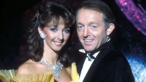 Debbie McGee has curly brown hair and is wearing large diamond-type earring and a necklace. The photo shows the folded upturned yellow edging of her dress. She is standing face-to-face with Paul Daniels, who has a swept over hair style and is wearing a tuxedo.
