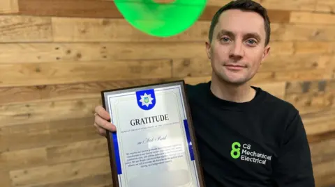 BBC Nick Reid seated in a meeting room holding a plaque from a Ukrainian police force. The plaque is silver and Nick is wearing a black polo with the C8 company logo. Nick is a white male with brown hair, blue eyes and appears to be in his thirties. 