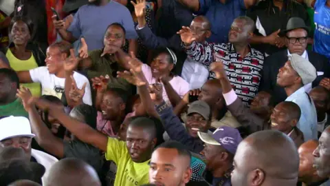 A shot of a crowd of people at a public meeting. Many are smiling and waving their arms in the air.