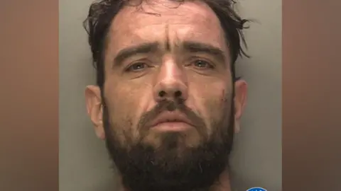 A mugshot of a man with dark hair and a dark beard. He is wearing a beige t-shirt and has his chin tilted upward.