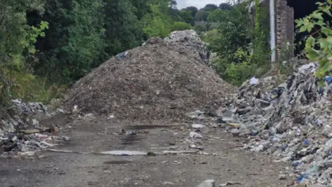UGC A large mound of waste has been piled next to a derelict building missing parts of a wall. The waste has been piled in lines on either side of the road up to the main pile.