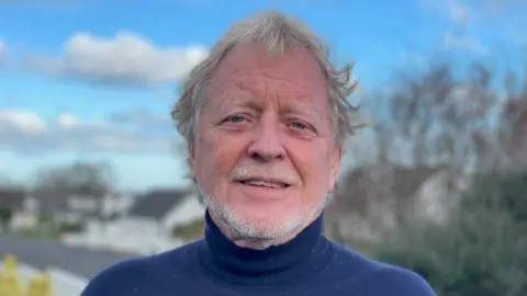 David Latimer. He has grey wavy hair and shirt beard, wearing a blue polo neck. Tress and houses are blurred in the background.