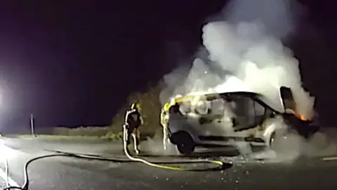 A burning van with a firefighter standing next to it with a hose.