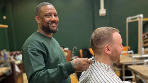 Jae Walters in a green jumper cuts a man's hair in the barbershop.
