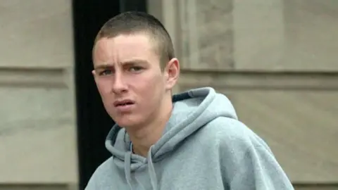 a young man with cropped brown hair and wearing a grey hoodie is standing outside a court building